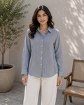 A woman stands outdoors wearing a light blue and white vertically striped button-up shirt with white pants. The background features a neutral beige wall with leaf shadows, a potted tree on the left, and a wooden chair on the right.