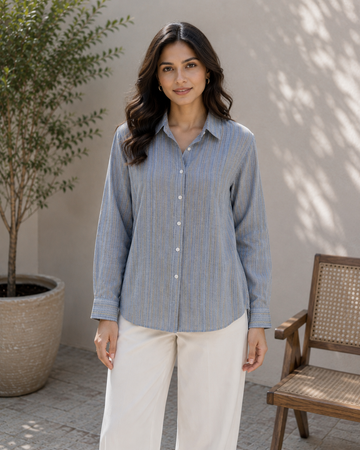 A woman stands outdoors wearing a light blue and white vertically striped button-up shirt with white pants. The background features a neutral beige wall with leaf shadows, a potted tree on the left, and a wooden chair on the right.