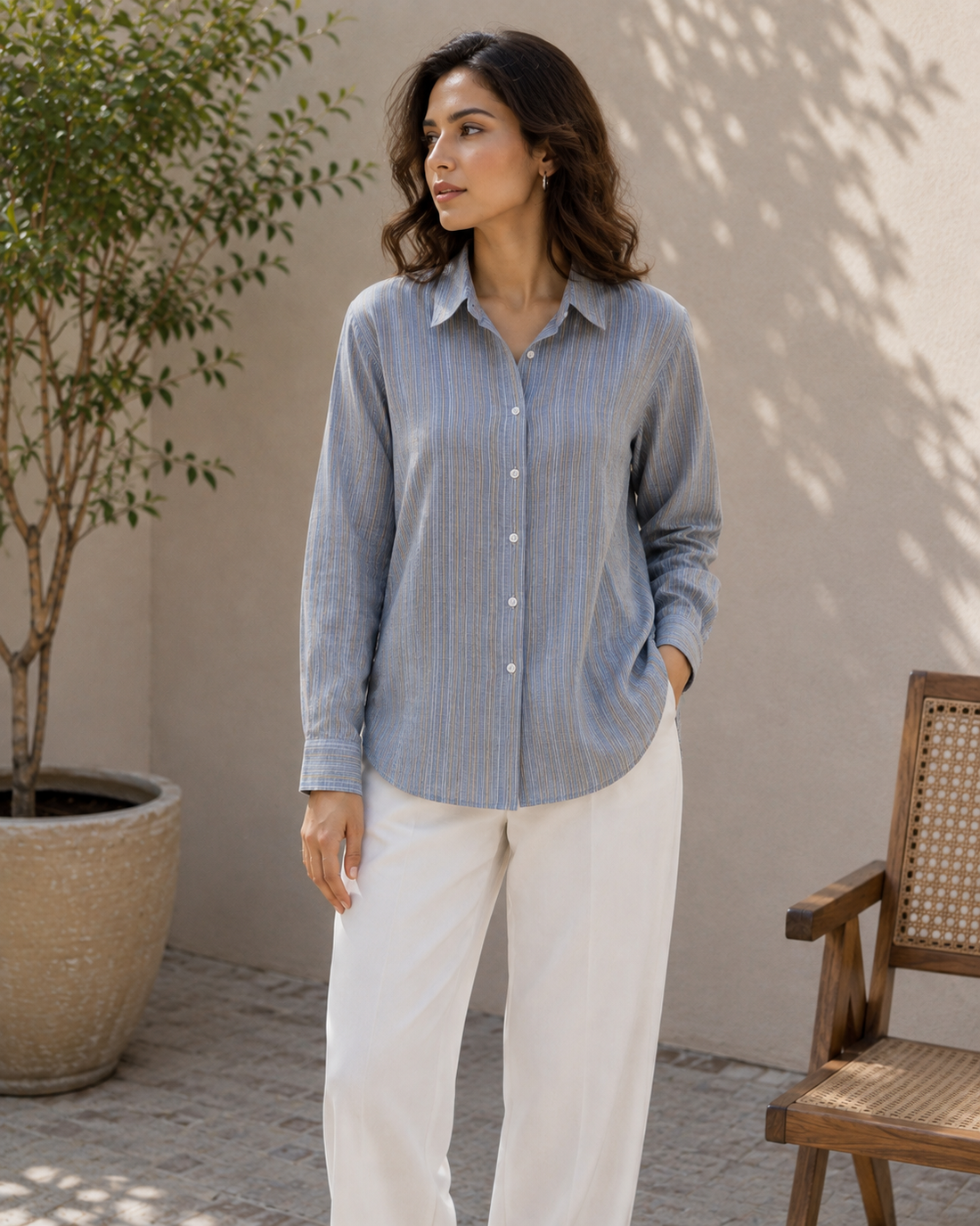 A woman stands outdoors wearing a light blue and white vertically striped button-up shirt with white pants. The background features a neutral beige wall with leaf shadows, a potted tree on the left, and a wooden chair on the right.