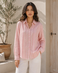 A woman wearing a light pink and white vertically striped long-sleeve button-up shirt with white pants, standing in a sunlit indoor-outdoor setting with a potted olive tree and a wooden door in the background.