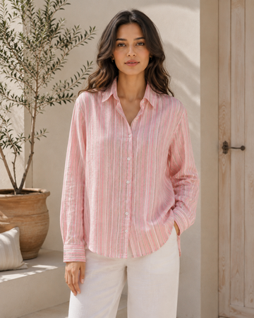 A woman wearing a light pink and white vertically striped long-sleeve button-up shirt with white pants, standing in a sunlit indoor-outdoor setting with a potted olive tree and a wooden door in the background.