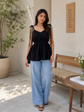 A woman modeling a black sleeveless peplum top with sheer shoulder straps and side cutouts, paired with light blue wide-leg jeans, standing in a sunlit indoor-outdoor space with a bench, table, and potted plant.