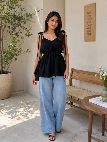 A woman modeling a black sleeveless peplum top with sheer shoulder straps and side cutouts, paired with light blue wide-leg jeans, standing in a sunlit indoor-outdoor space with a bench, table, and potted plant.