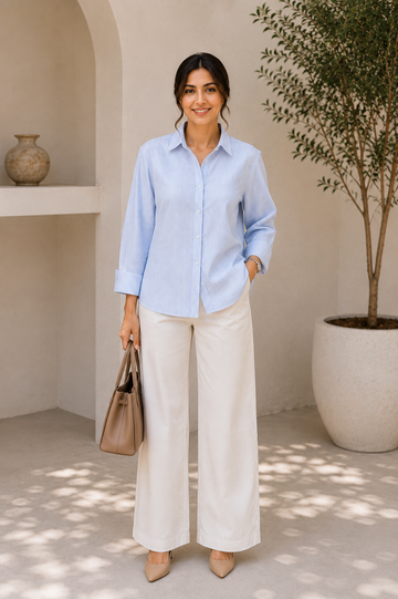 A woman stands in a sunlit architectural setting wearing a light blue button-up shirt and wide-leg cream pants, holding a taupe handbag.