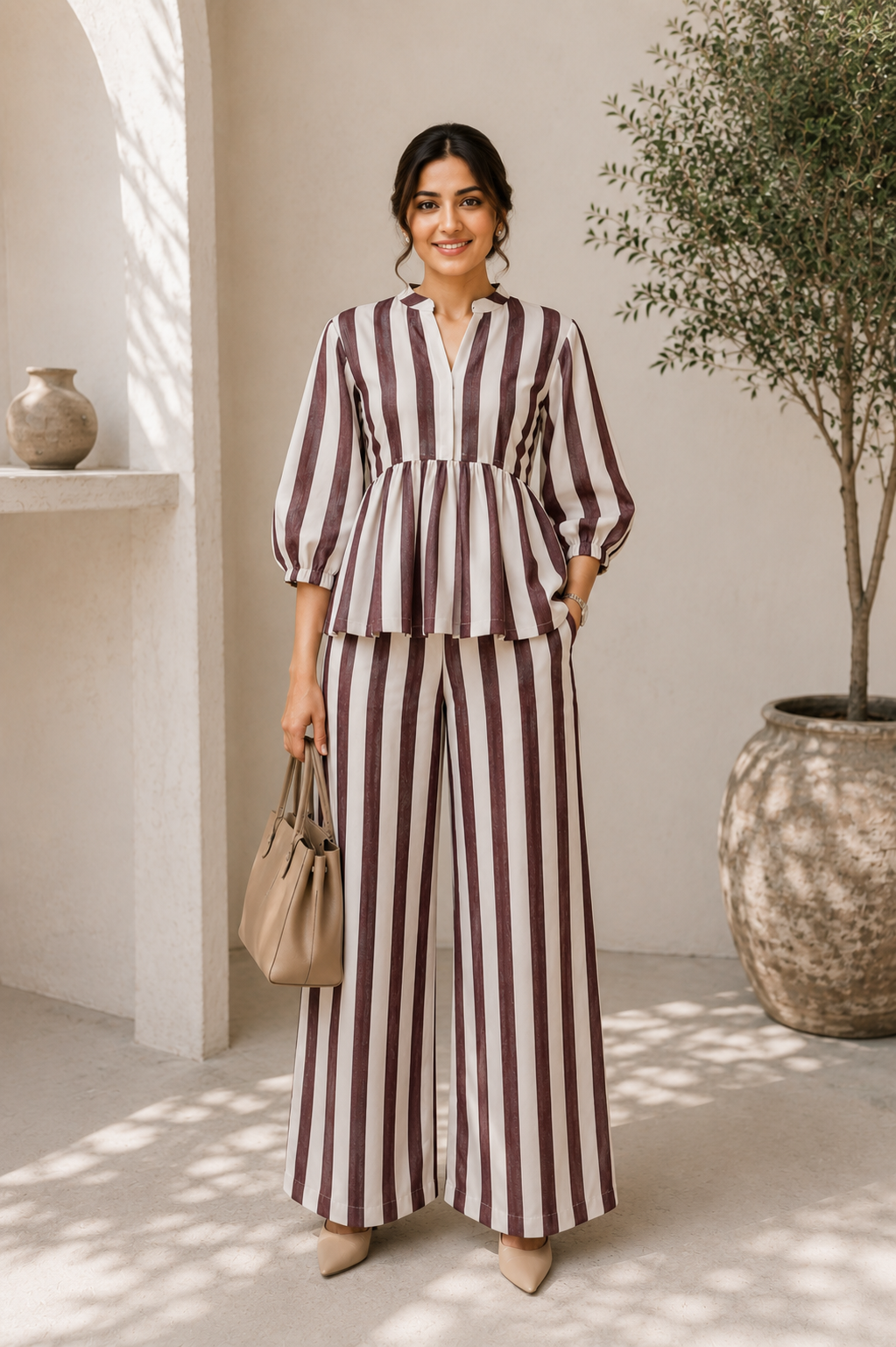A woman models a matching two-piece set featuring vertical burgundy and cream stripes, consisting of a peplum-style top with three-quarter sleeves and wide-leg trousers. She is standing in a sunlit, modern architectural setting with a potted tree and a small vase on a shelf.