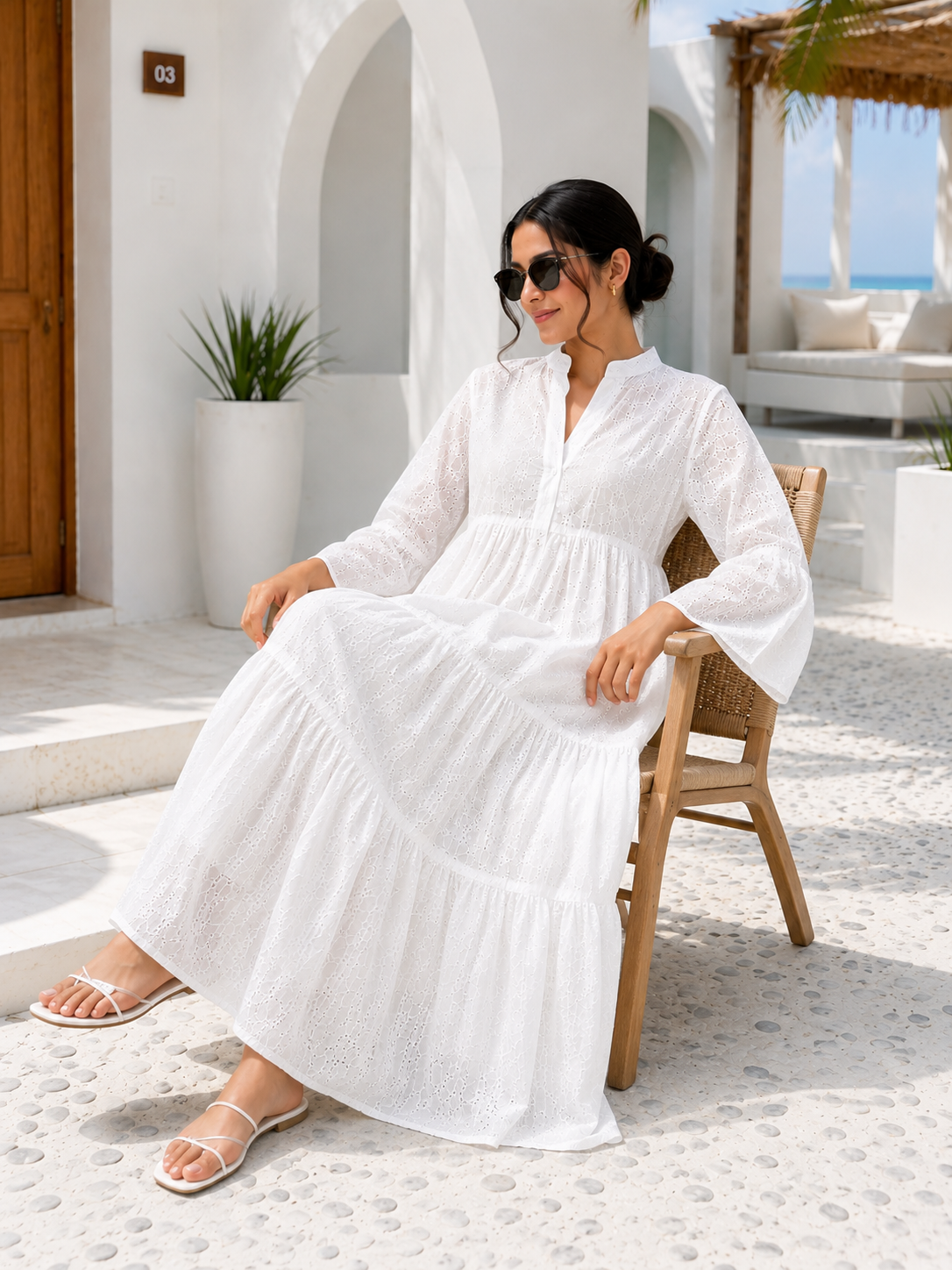 A woman wearing a white tiered maxi dress with three-quarter sleeves and a split neckline, walking outdoors in a sunny, modern Mediterranean-style courtyard. She is holding a woven straw handbag.