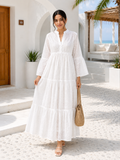 A woman wearing a white tiered maxi dress with three-quarter sleeves and a split neckline, walking outdoors in a sunny, modern Mediterranean-style courtyard. She is holding a woven straw handbag.