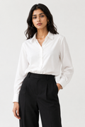 A woman modeling a white long-sleeve button-up shirt tucked into black high-waisted trousers, photographed against a clean light gray studio background.